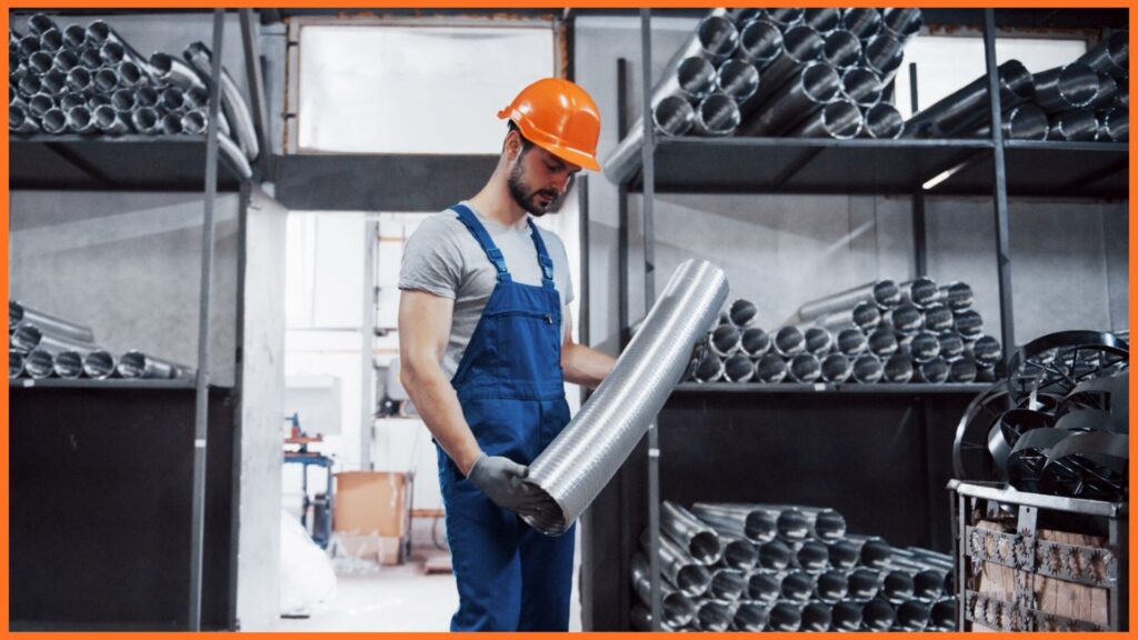 Factory worker in hard hat inspecting a roll of flexible metal ducting in an industrial warehouse