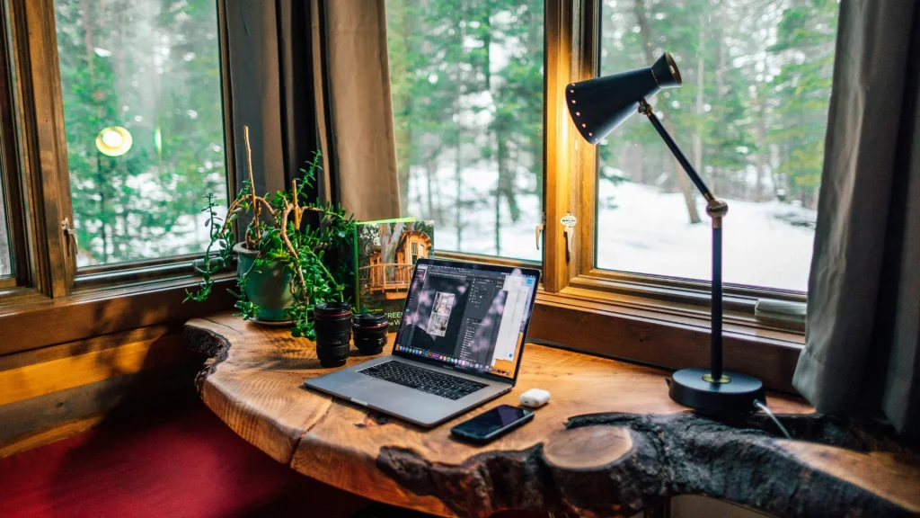 Cozy rustic live-edge wooden desk setup with a laptop, lamp, and potted plant by a window overlooking a snowy forest landscape