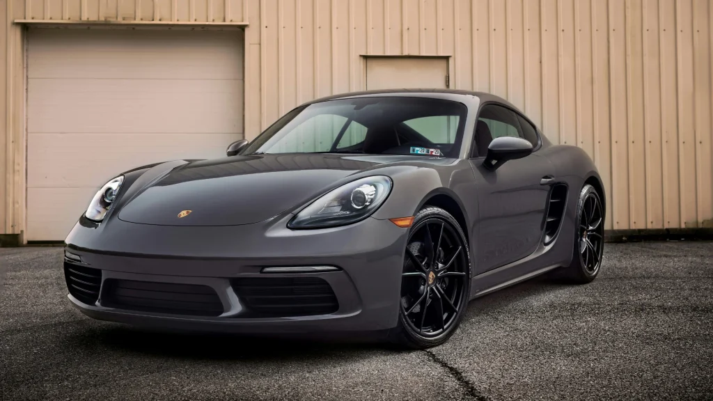 Sleek dark grey Porsche 718 Cayman sports coupe with black wheels parked outdoors in front of a neutral-colored building